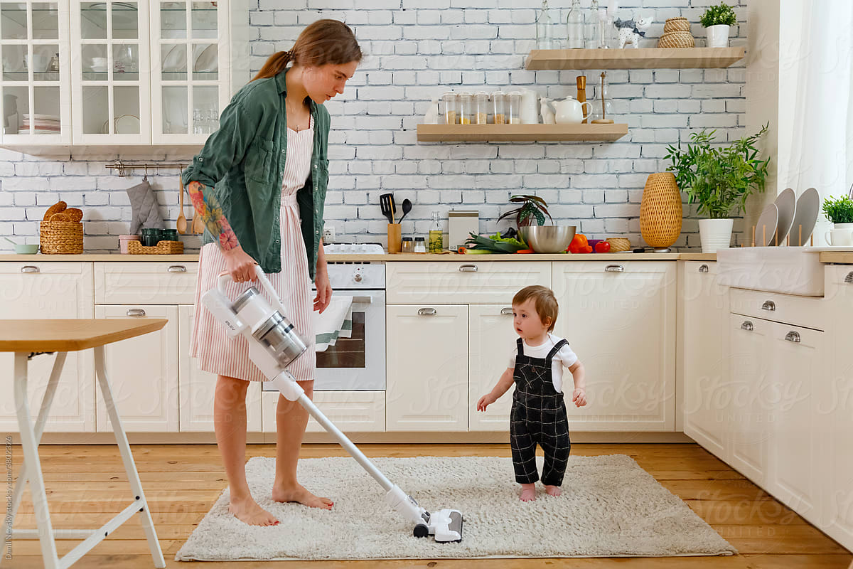 Focused housewife vacuum cleaning floor in kitchen near cute toddler son
