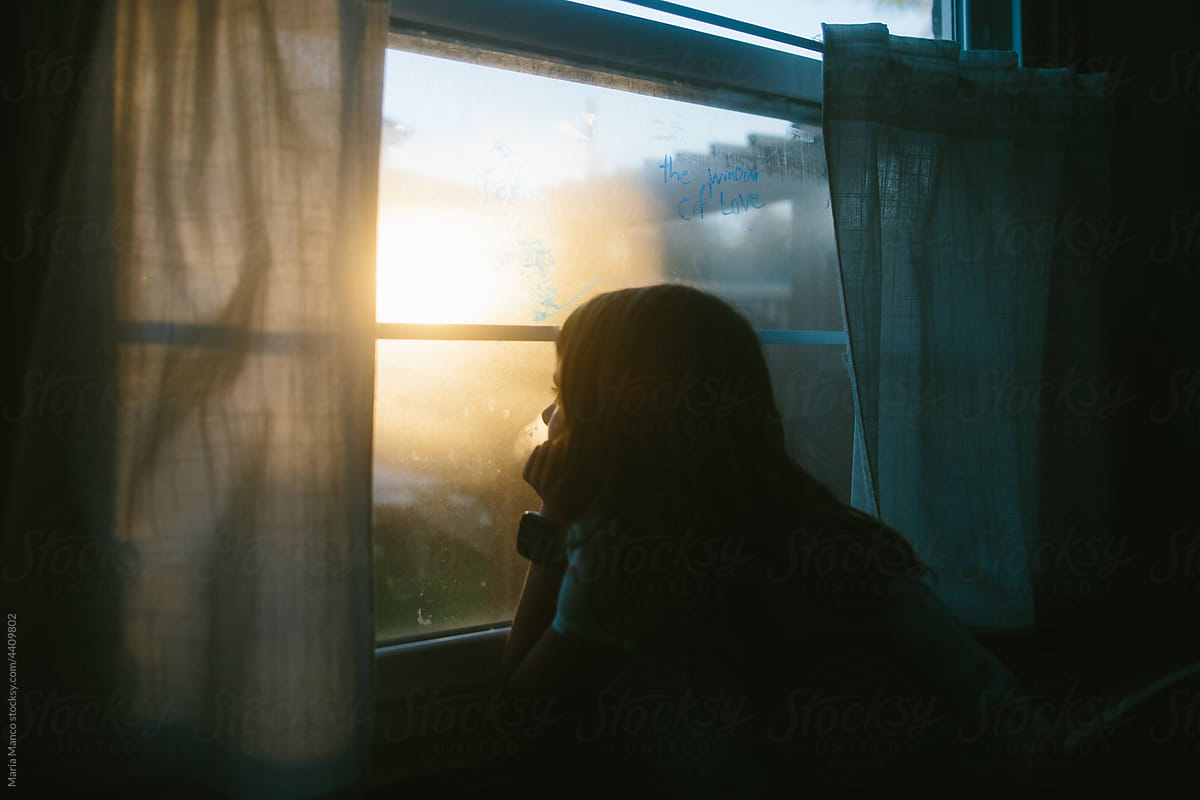 Young girls looks out window with sun setting outside