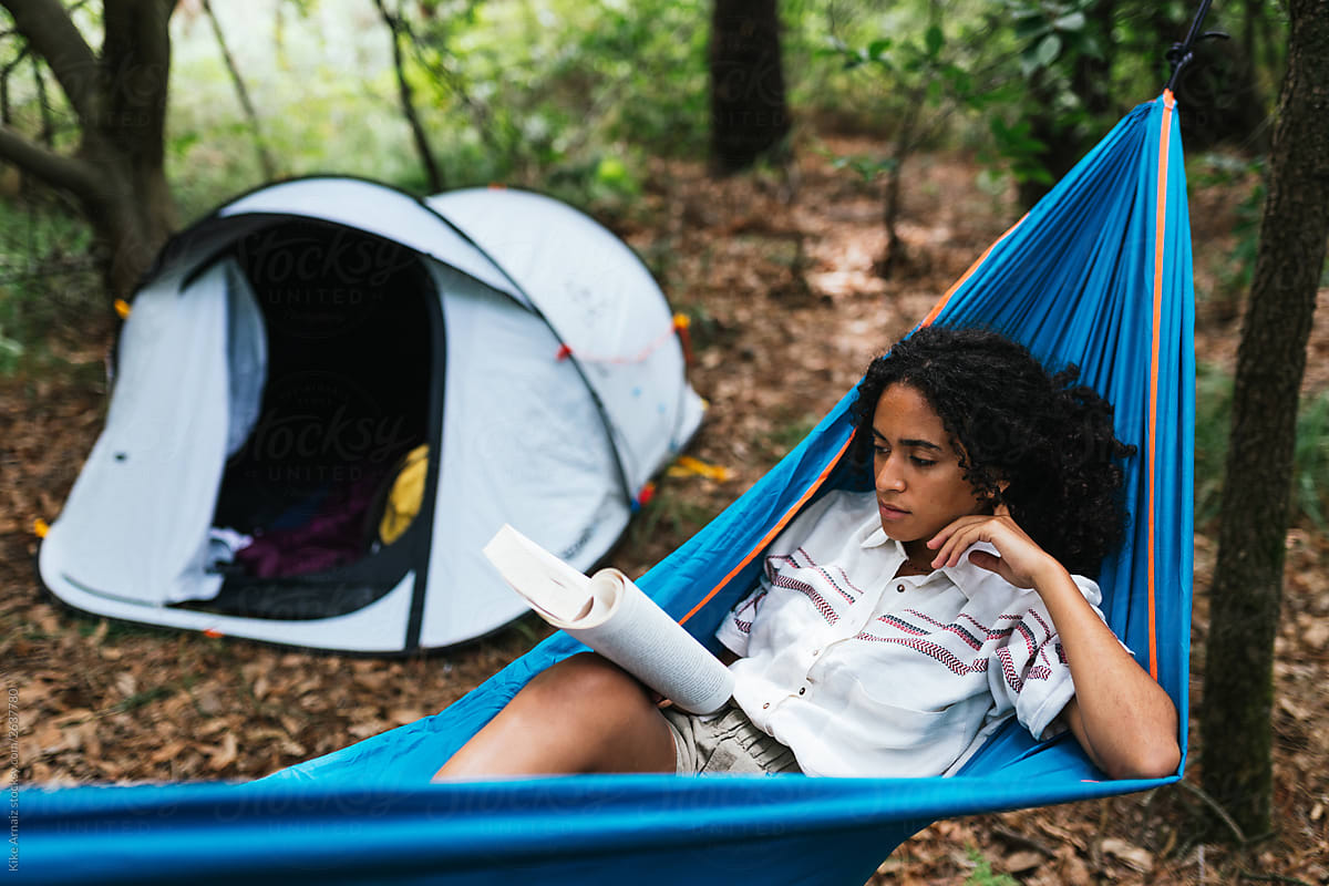 african american woman lying down on a hammock reading a book for camping