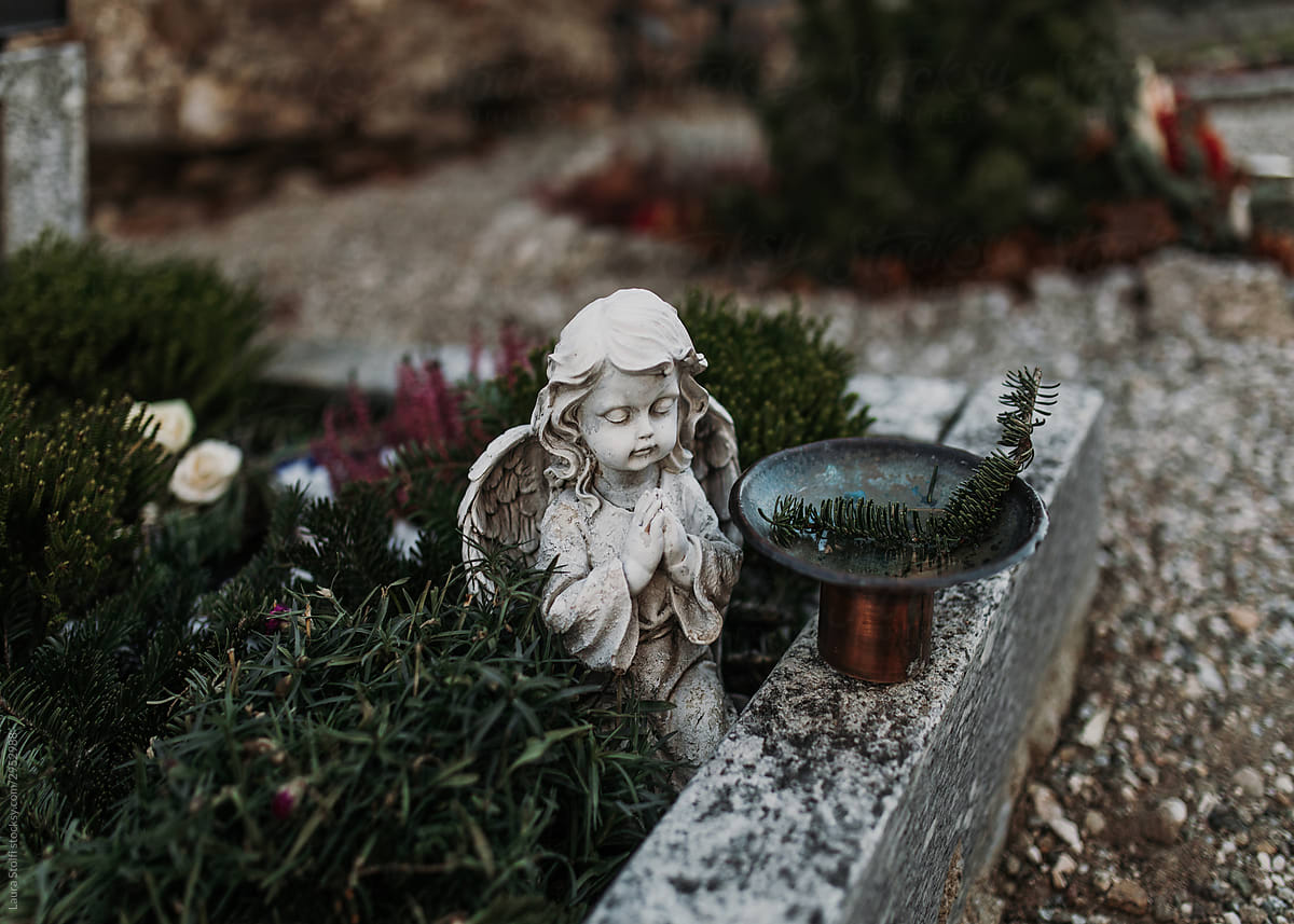 Serene cherub angel statue made with concrete praying on a grave decorated with flowers and plants