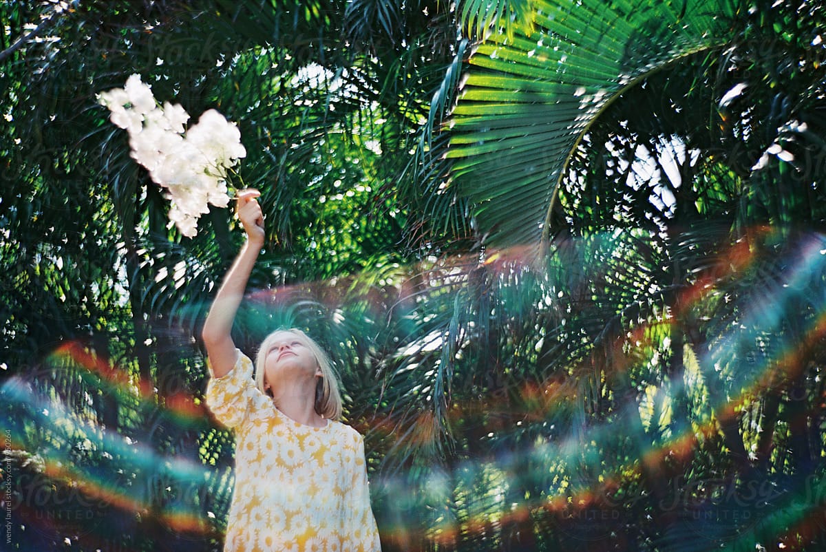 girl in yellow dress holding white flowers against green palms and rainbow flare