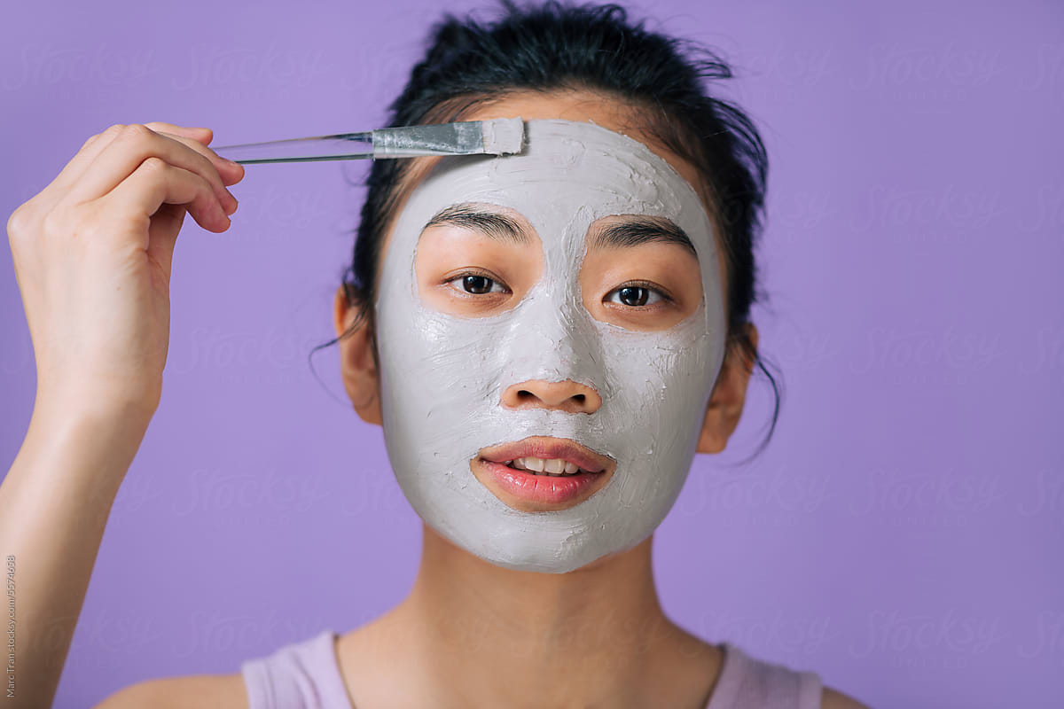 A woman is applying a clay mask to her face, using brush accessory.