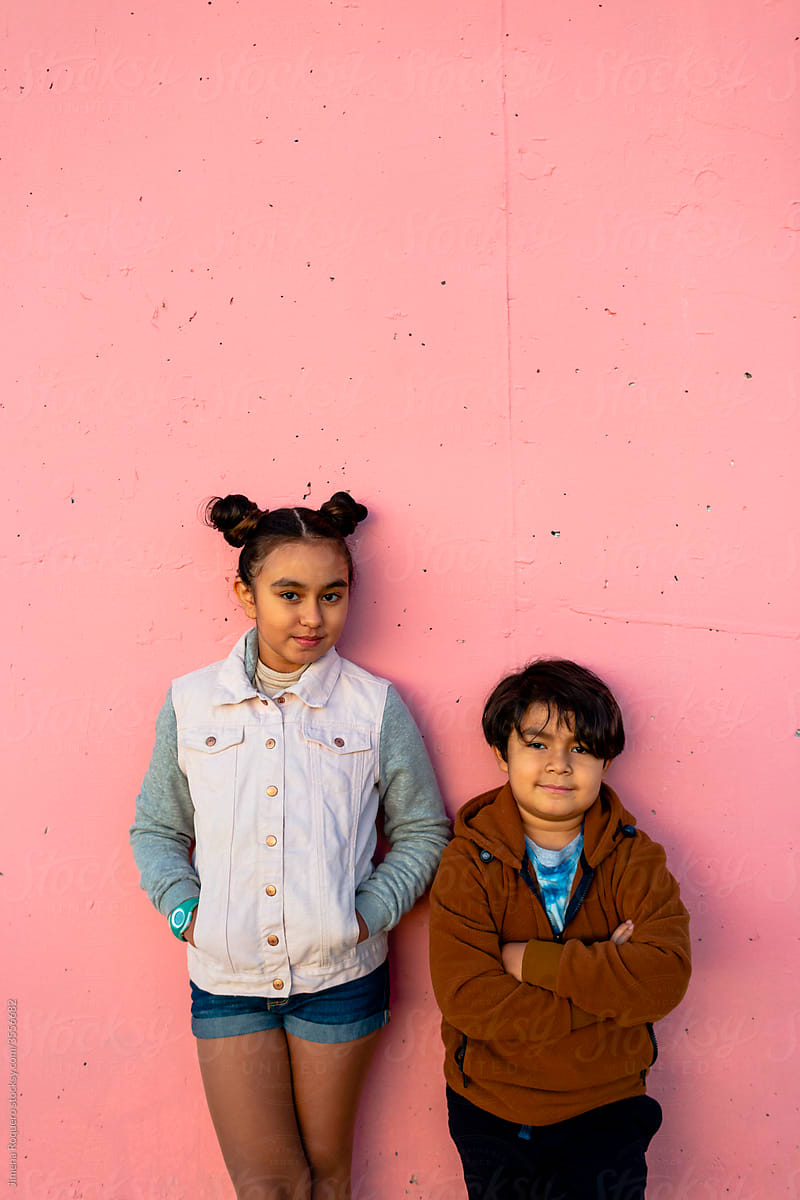 Hispanic Brother and sister posing over pink wall looking at camera.