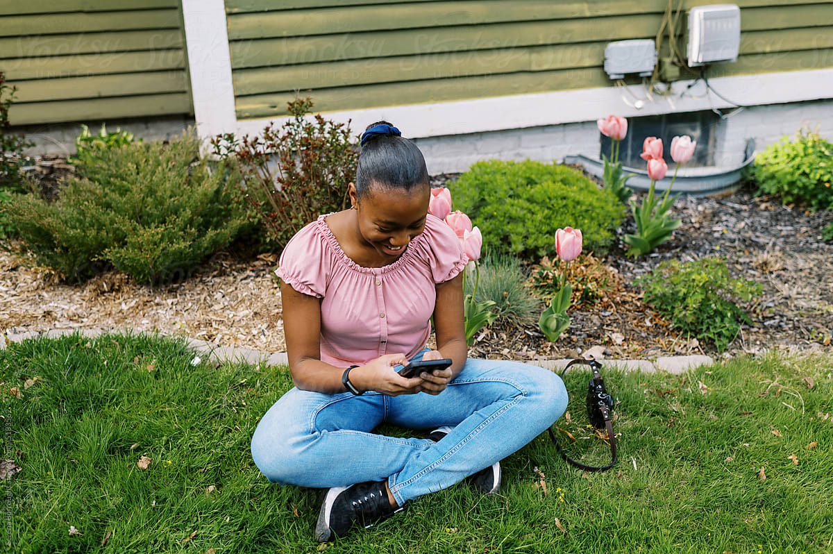 smiling black girl sitting down in grass with her phone and camera
