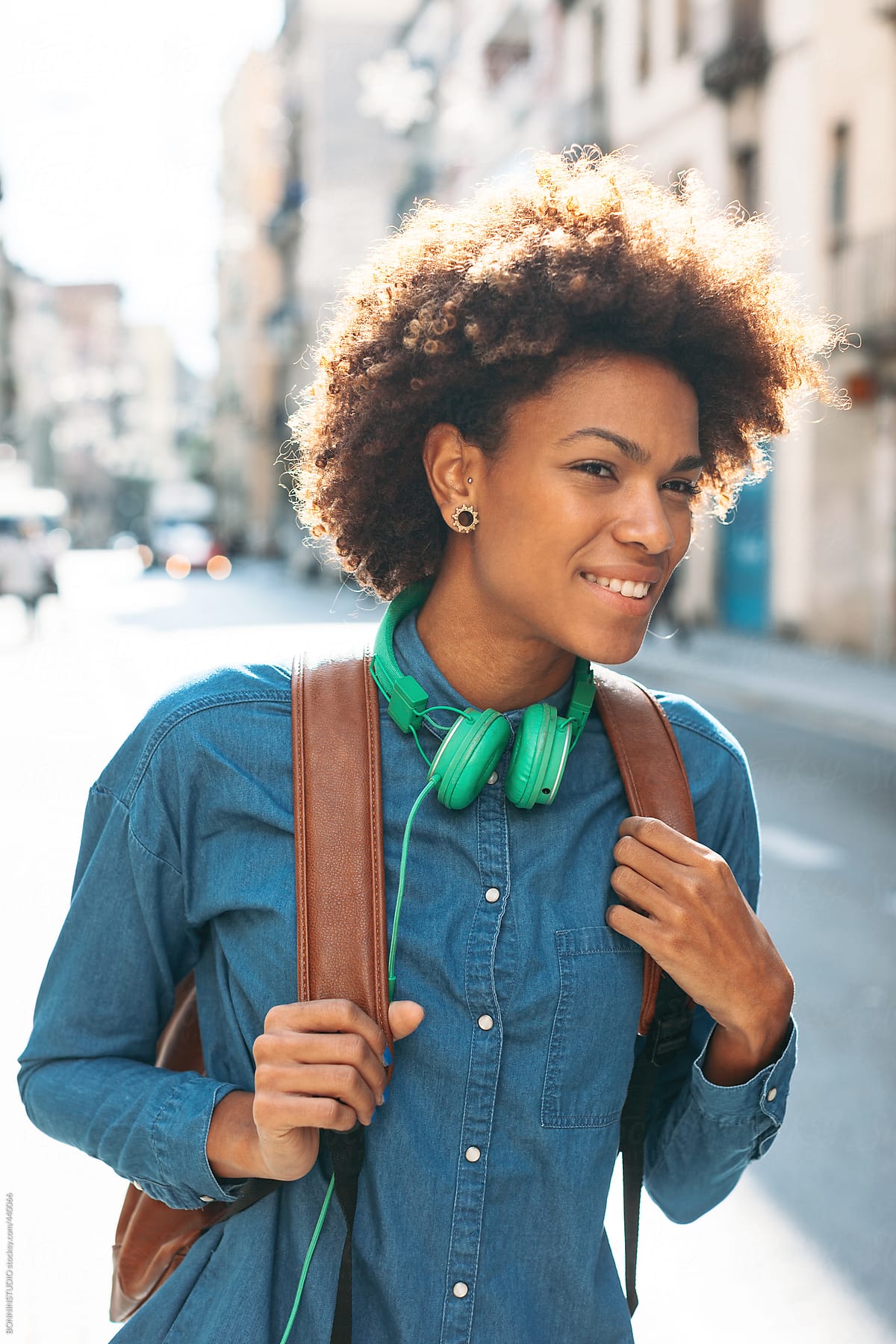 Afro american woman with leather backpack and green headphones smiling in outdoors.