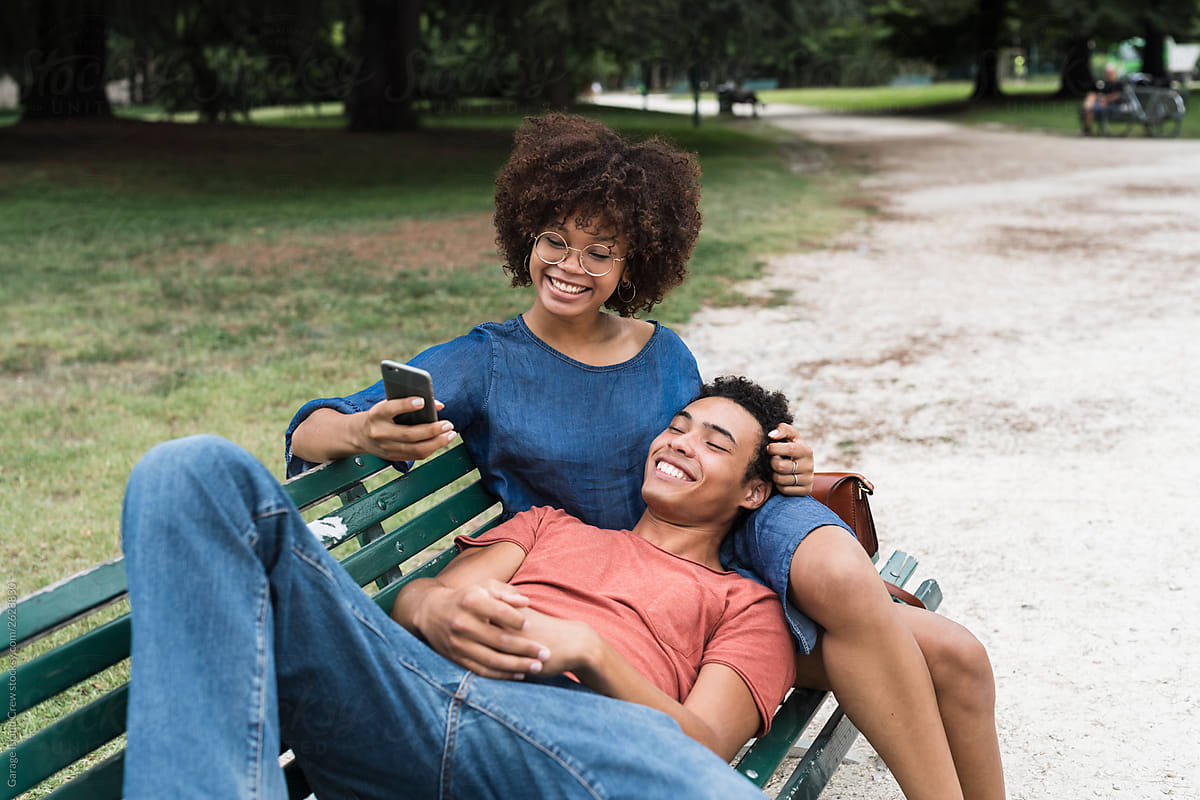 Happy young black couple sitting on the bench at the park using