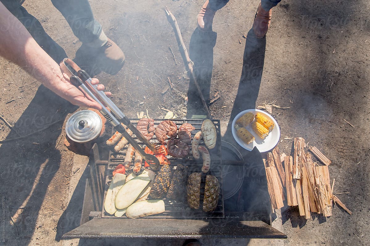 Overhead view of campfire barbecue with grey nomad couple tending meat, vegetables and fruit cooking on grill