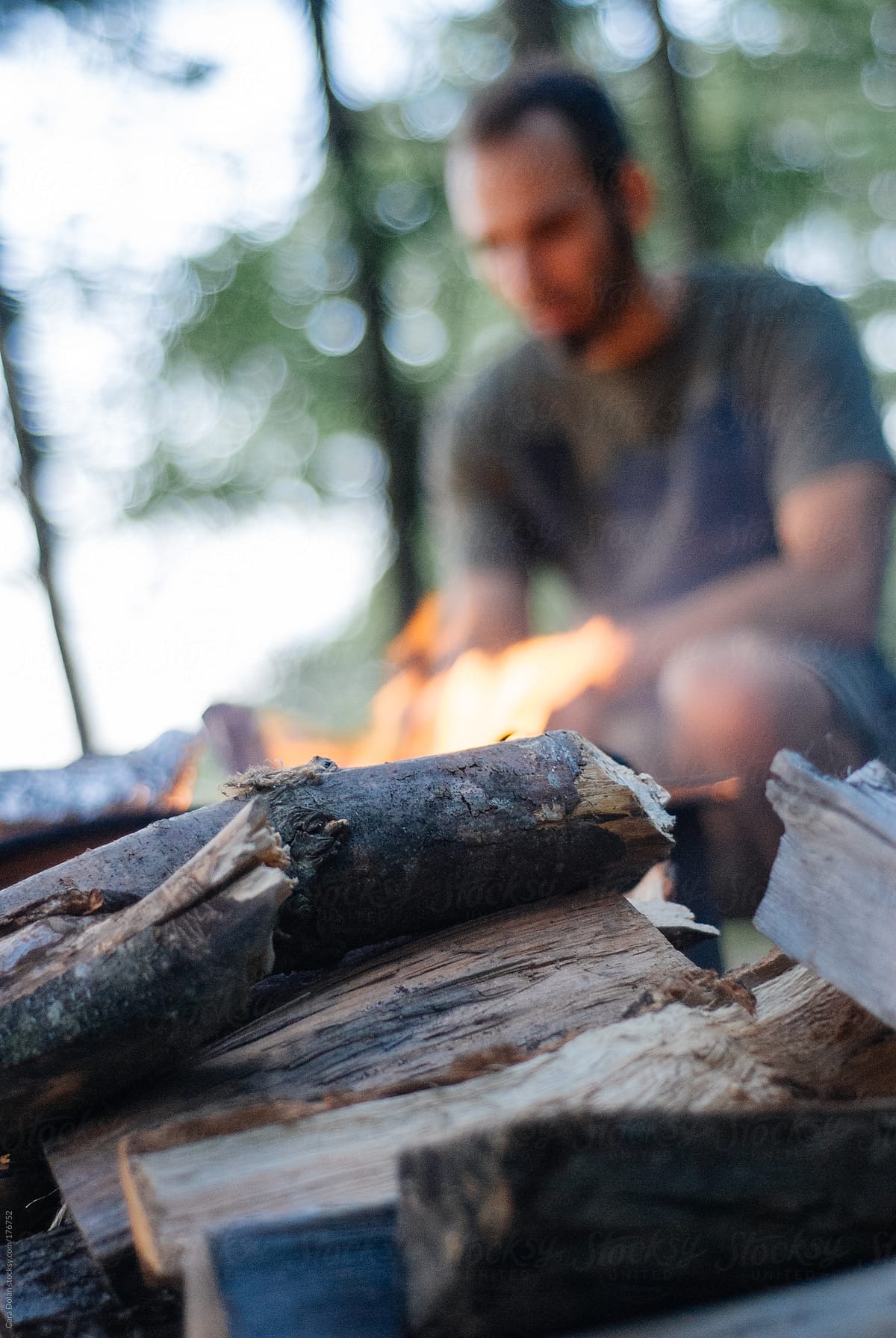 Man sits tending to a campfire, with firewood in foreground
