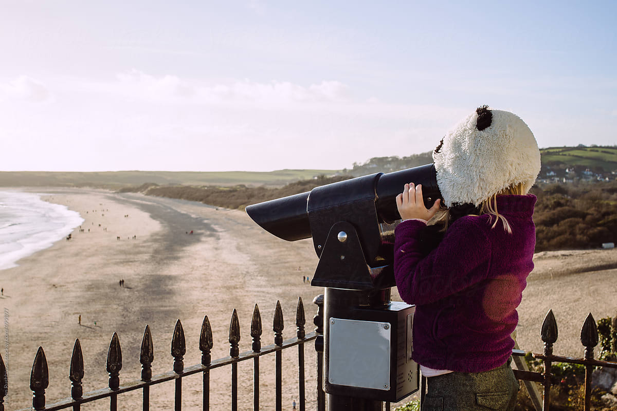 A little girl in a panda hat looking through a telescope overlooking a beach.