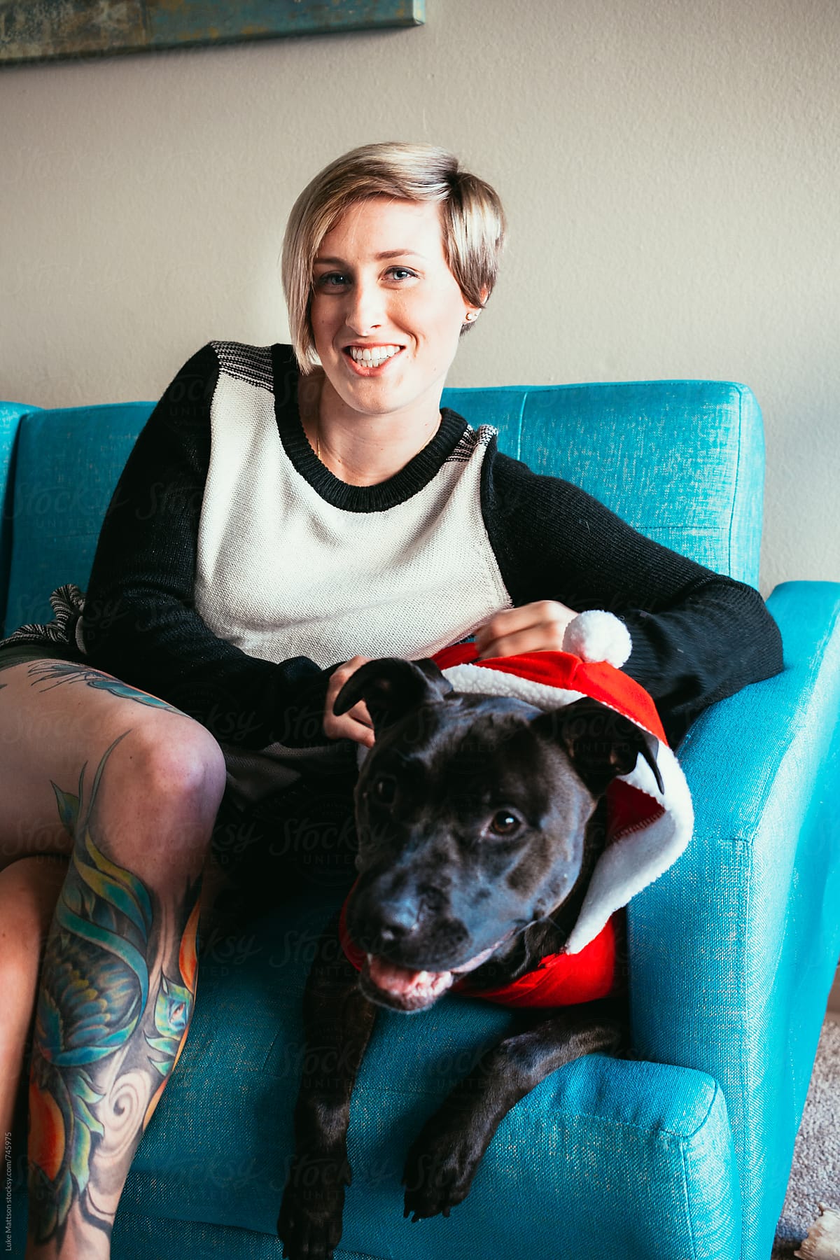 Smiling Young Blonde Tattooed Woman Sitting On Couch With Pet Pit Bull In Santa Claus Costume