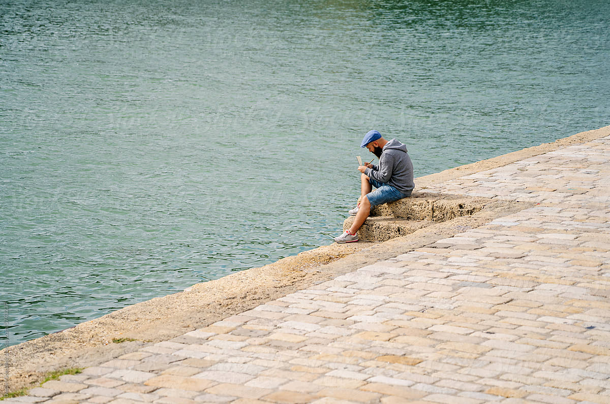Male sitting on waterfront of river and writing notes
