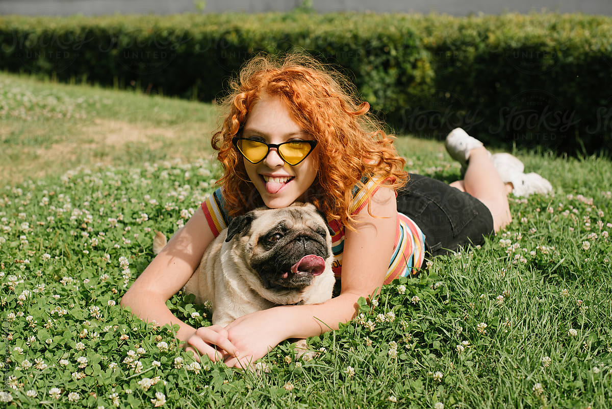 Ginger head in teh yellow glasses lies on the grass and holds her pug dog