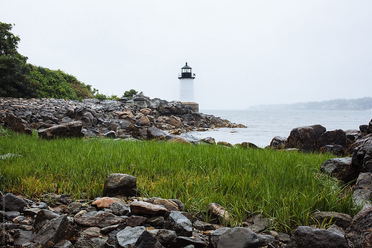A lighthouse in the distance with grass and rocks in the foreground. by J Danielle Wehunt for Stocksy United