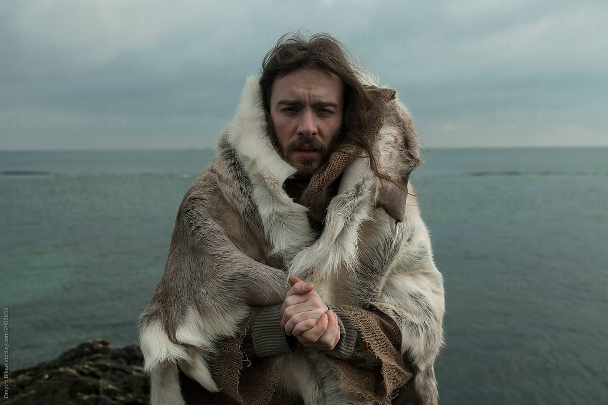 a guy with long hair and a beard against the backdrop of the sea in winter poses in animal skins