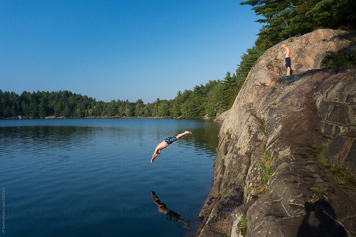 Man Gracefully Dives Headfirst Into Warm Summer Cottage Lake From Cliff Del Colaborador De