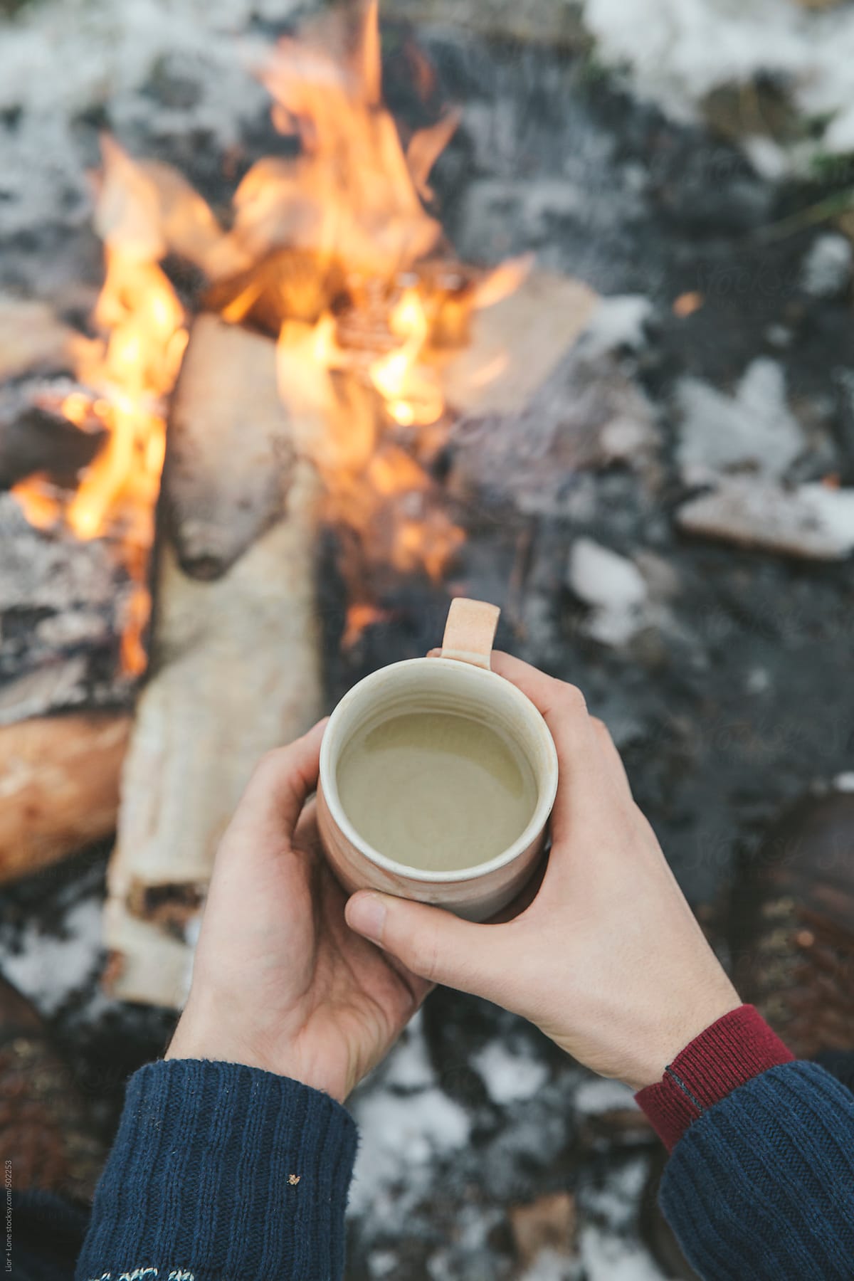 Man\'s hands holding a cup of tea next to bonfire outdoors