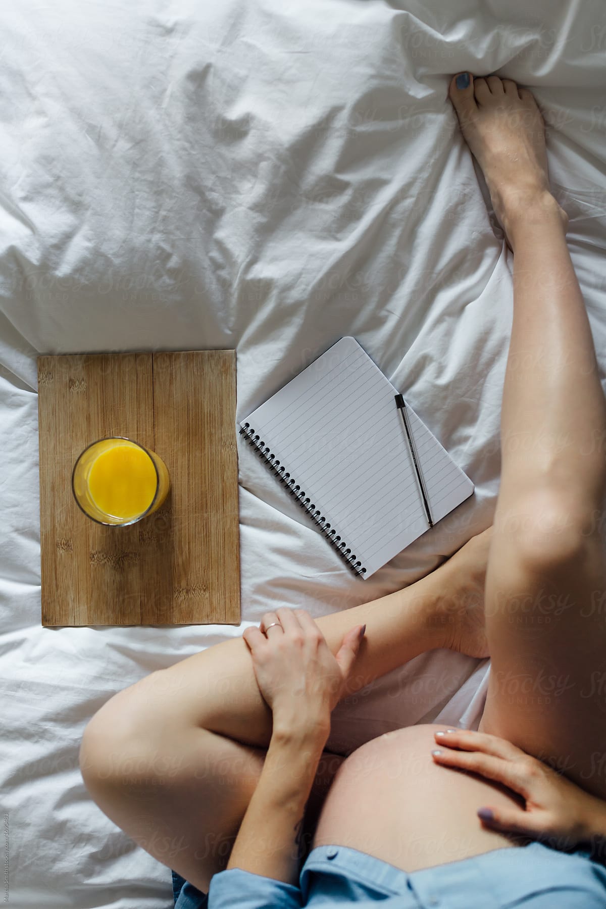 A pregnant women\'s bare legs and tummy from above. A glass of orange juice and a blank notebook are on the white sheeted bed.
