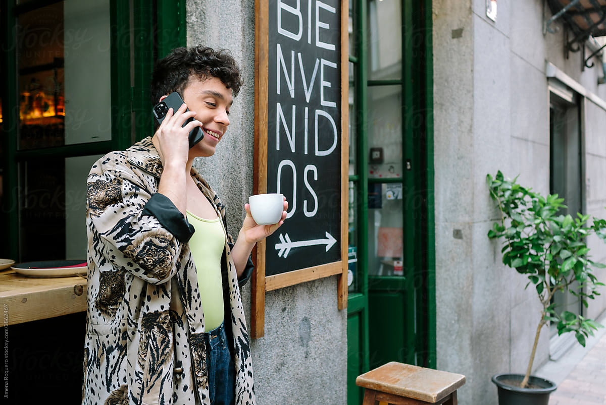 Young gay man outside of city café with smartphone drinking coffee and smiling