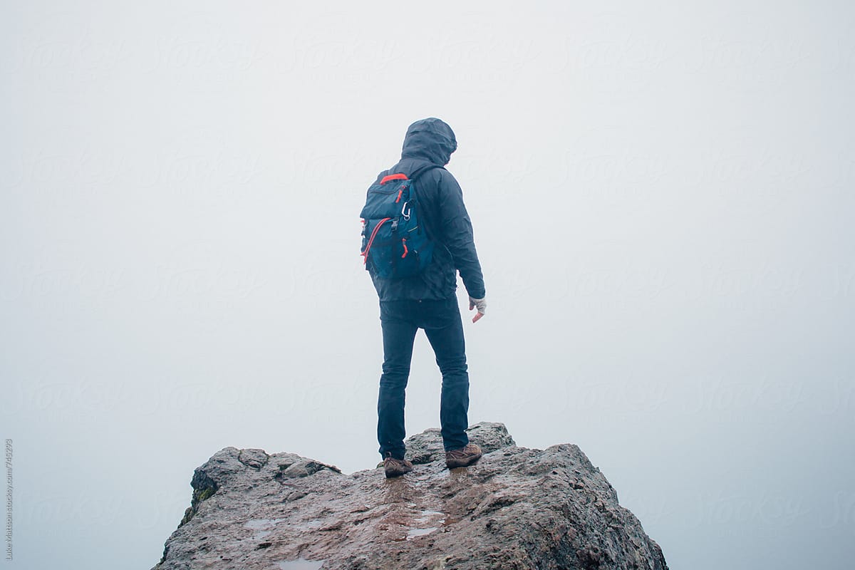 Hooded Hiker Wearing Backpack Stands On Rocky Ledge In Fog