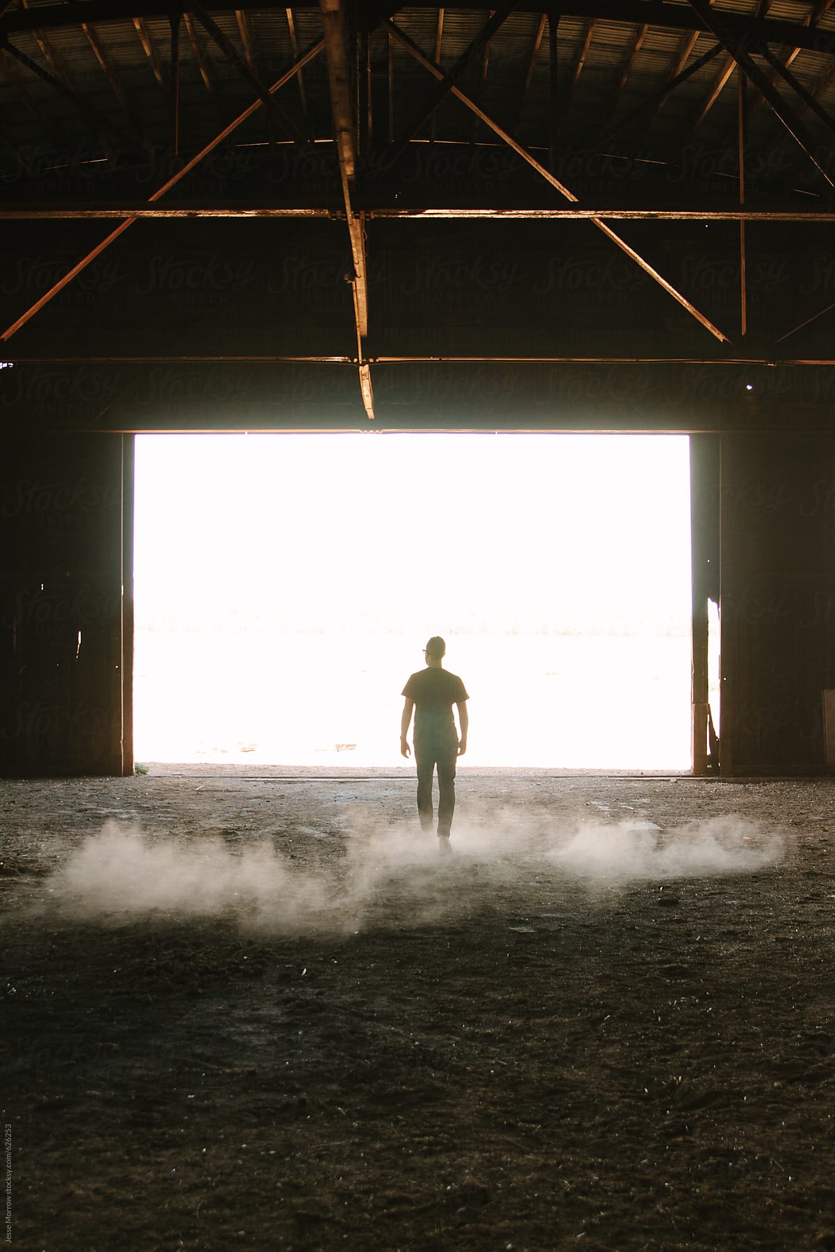 Young man walking through empty barn kicking up dirt floor with bright white behind silhouette