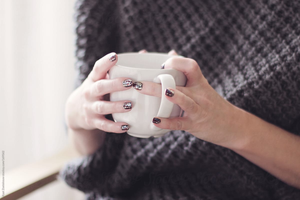 Close view of a woman with lacquered nails holding a white mug of coffee in her hands.