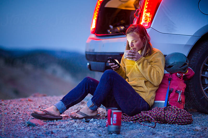 Young Adult Woman Uses Phone and Drinks Warm Beverage outside Car