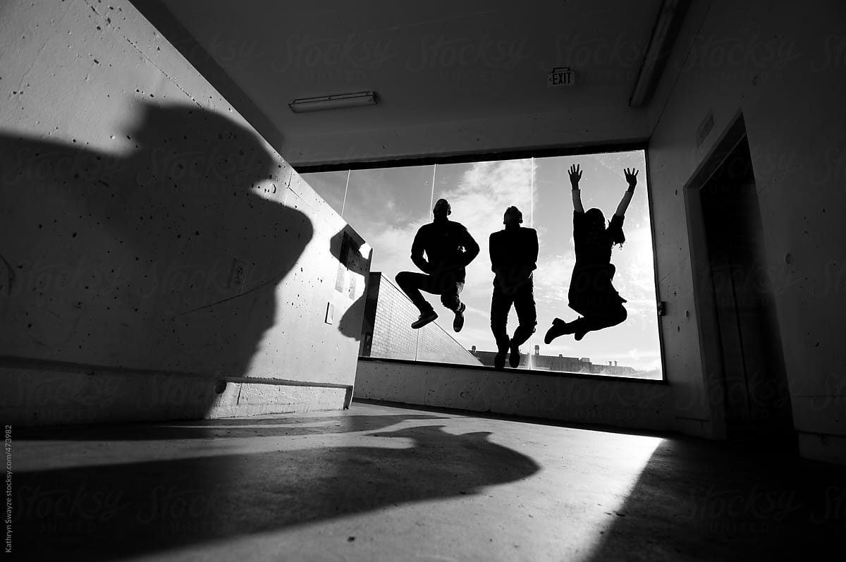 Three people jump in the stairwell of a parking garage, backlit by a bright window