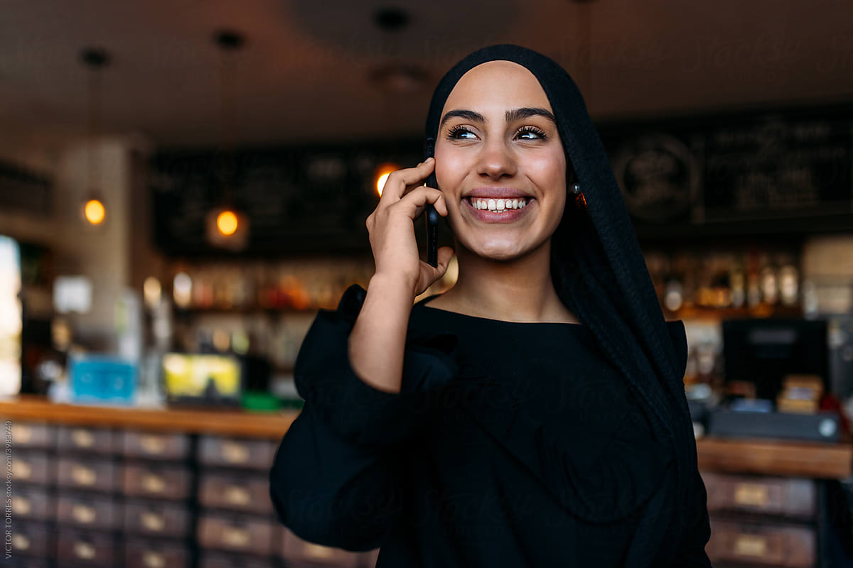 Cheerful stylish Muslim woman using cellphone in cafe