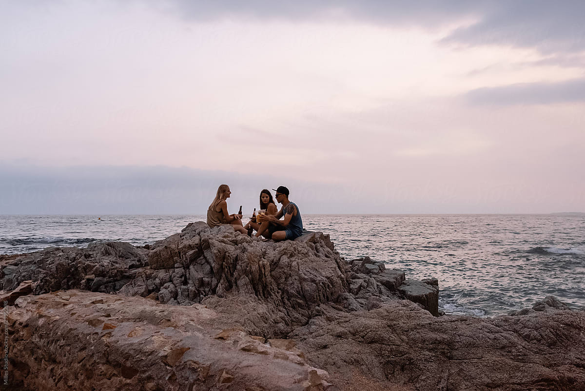 Friends drinking beer and communicating on stony seashore during sunset