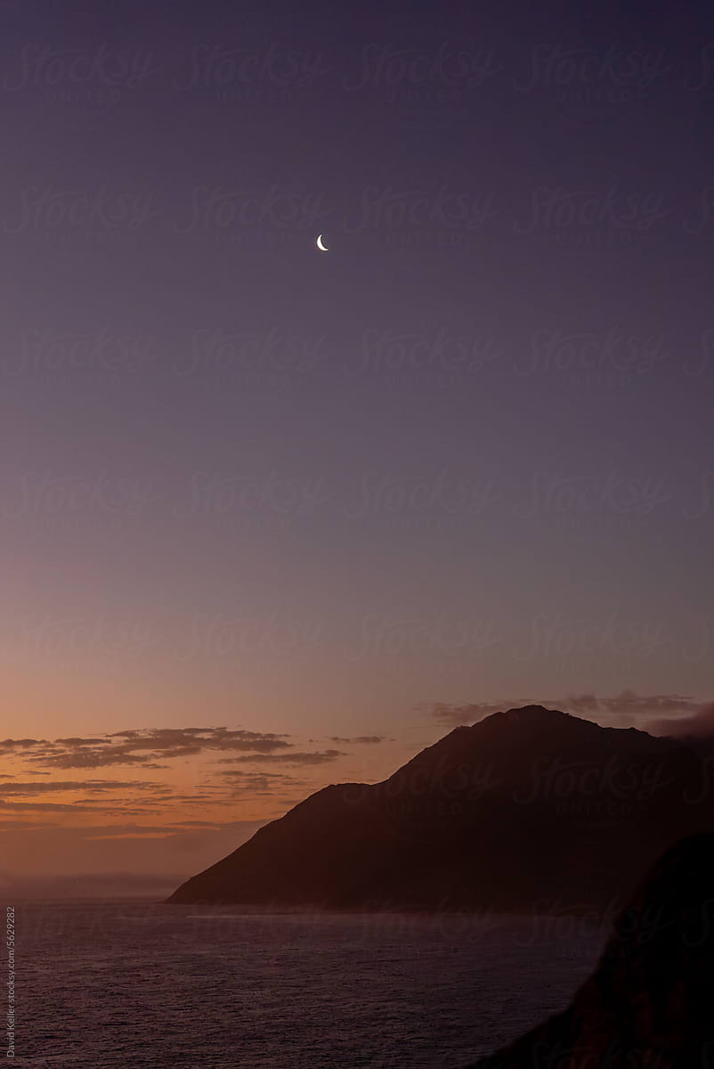 A sliver of the moon radiates above a cloudy sunset on the coast