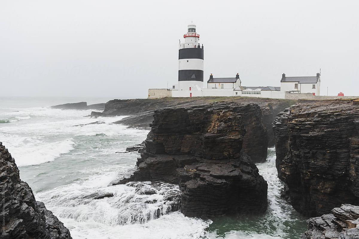Lighthouse in the fog with rocks and rough sea in the foreground.
