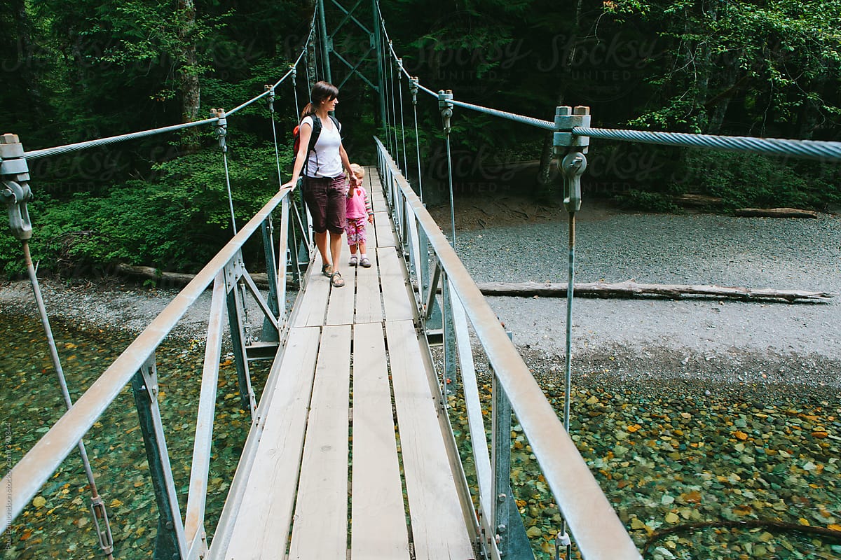 Mother and young daughter walking across suspension bridge and river