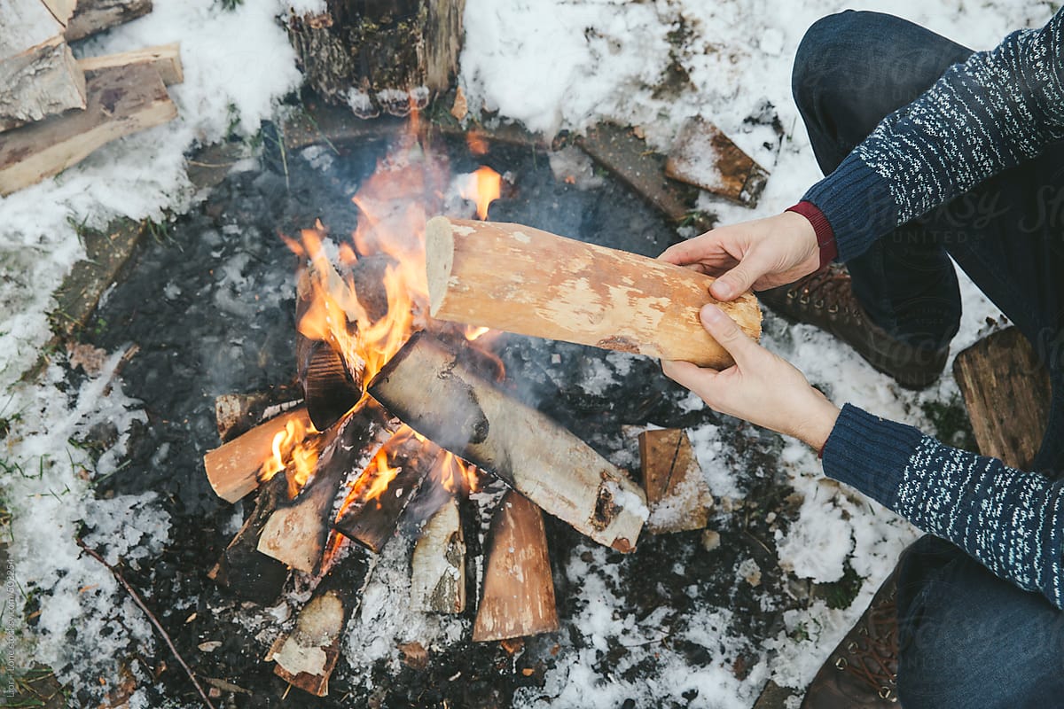 Hands addig wood to a bonfire outdoors in the snow