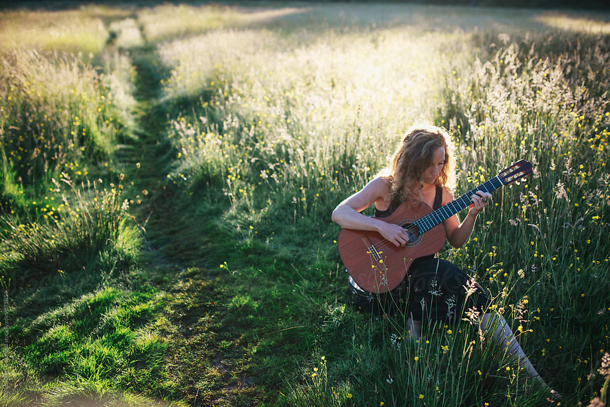 Young woman sitting in field at sunrise playing classical guitar and singing
