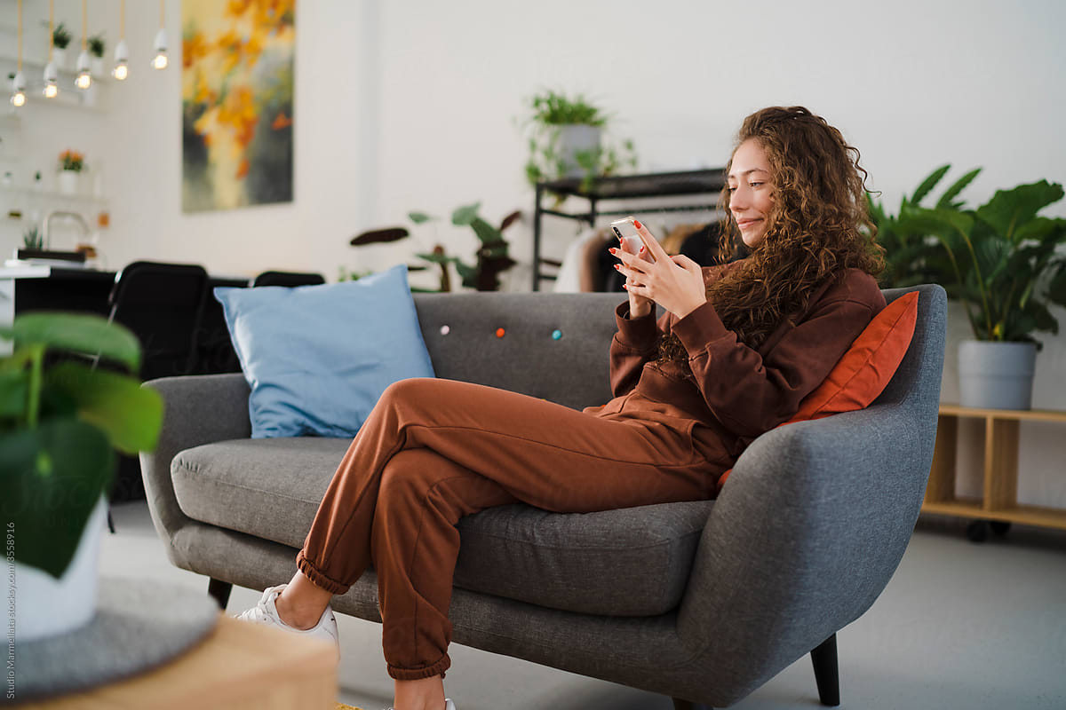 Happy female resting on couch with hot beverage and smartphone