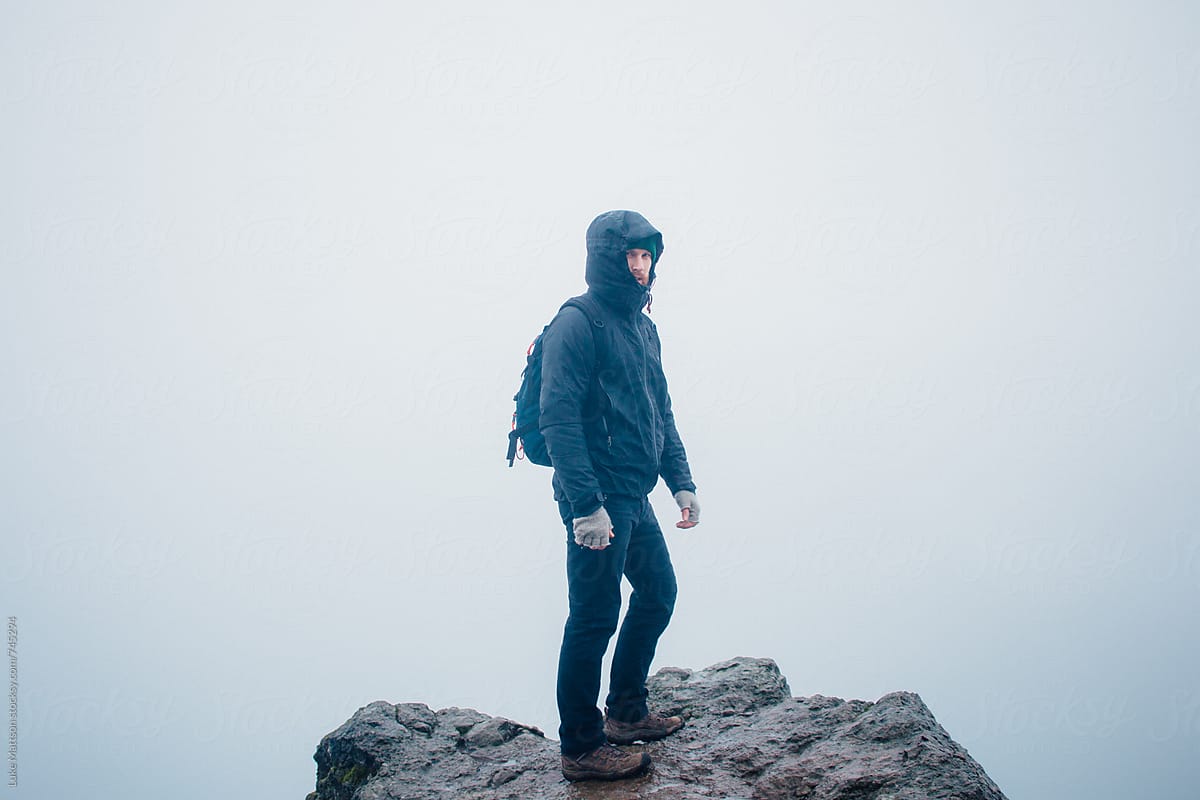 Hooded Hiker Wearing Backpack Standing On Rocky Ledge In Fog