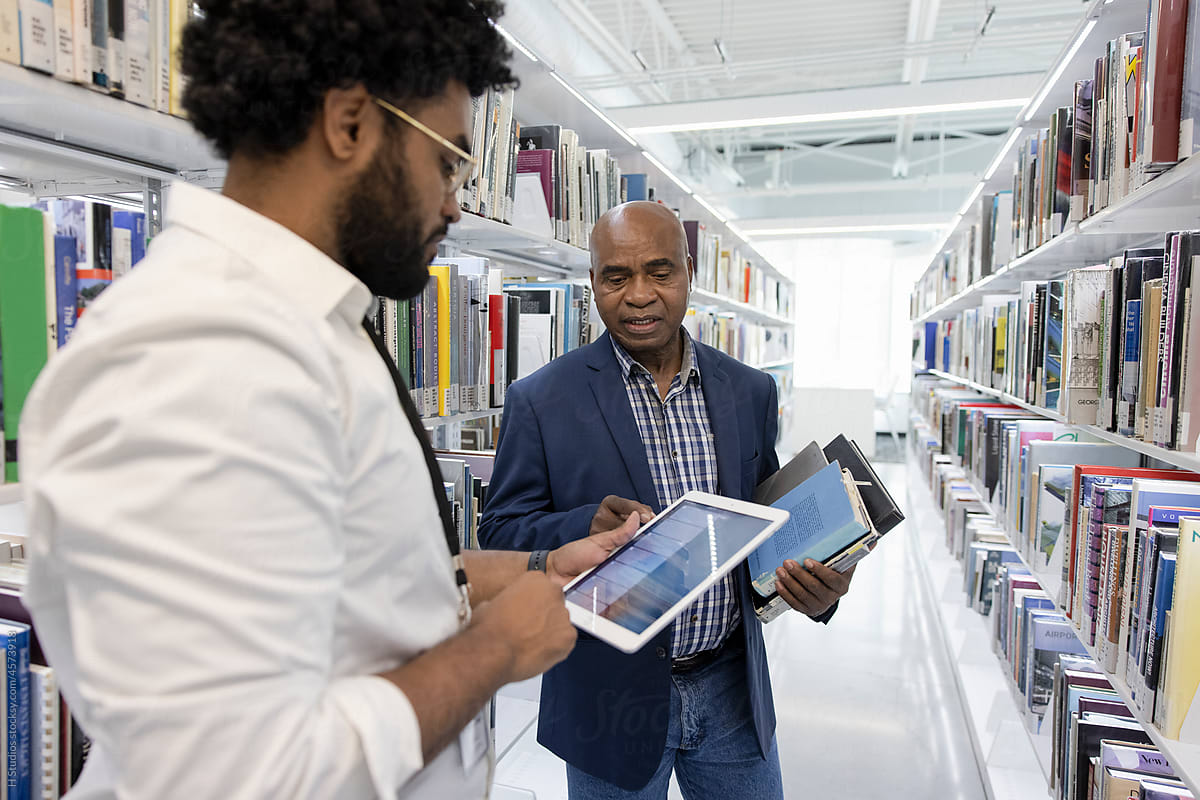 Librarian with digital tablet helping man with books in library.