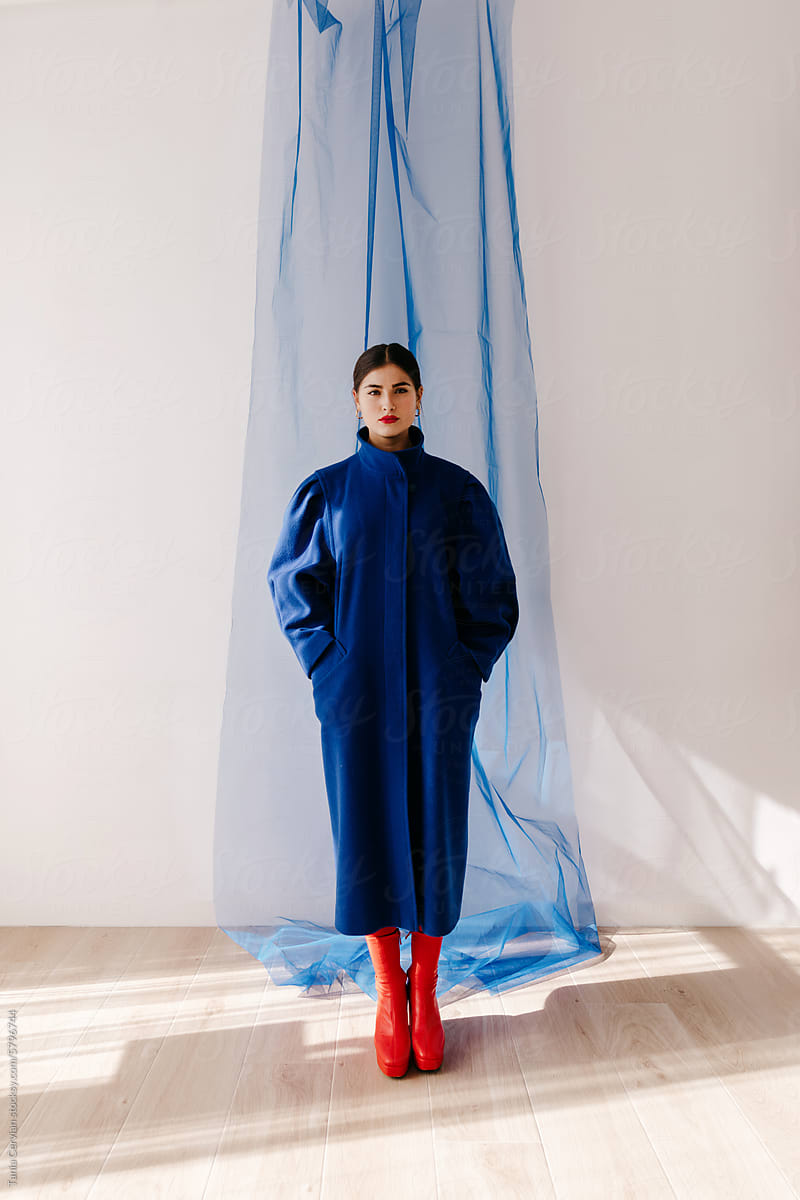 Stylish woman in coat standing against blue fabric in studio