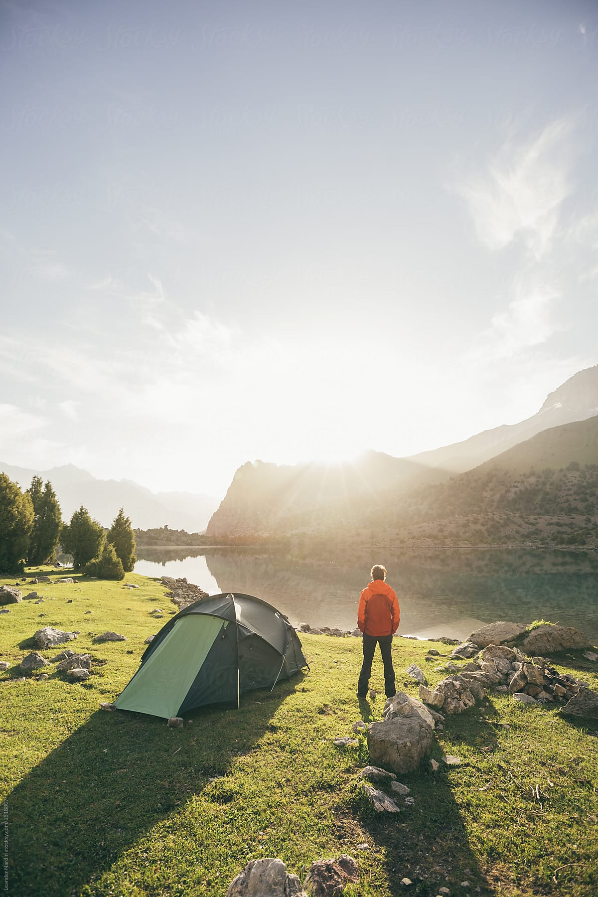 hiker in red jacket beside his tent enjoying sunrise at a mountain lake