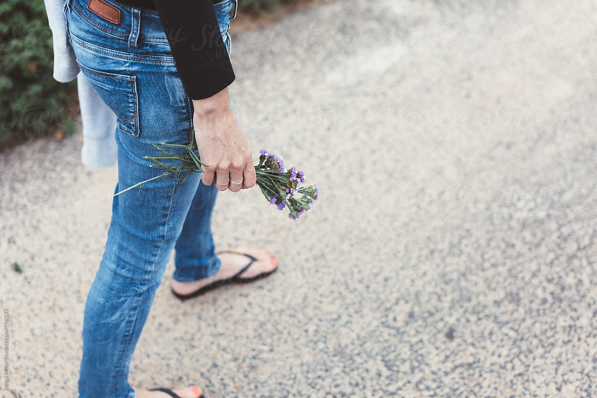Woman wearing blue denim pants is walking a long a road holding wild flowers in her hand
