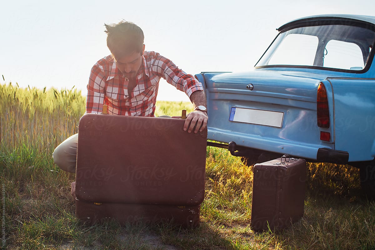 Retro-Styled Young Man Looking at Clothes in Vintage Leather Suitcase Next to Old Blue Car