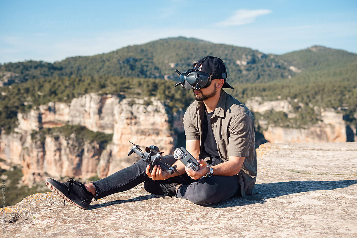 Bearded guy in VR goggles sitting on cliff with drone