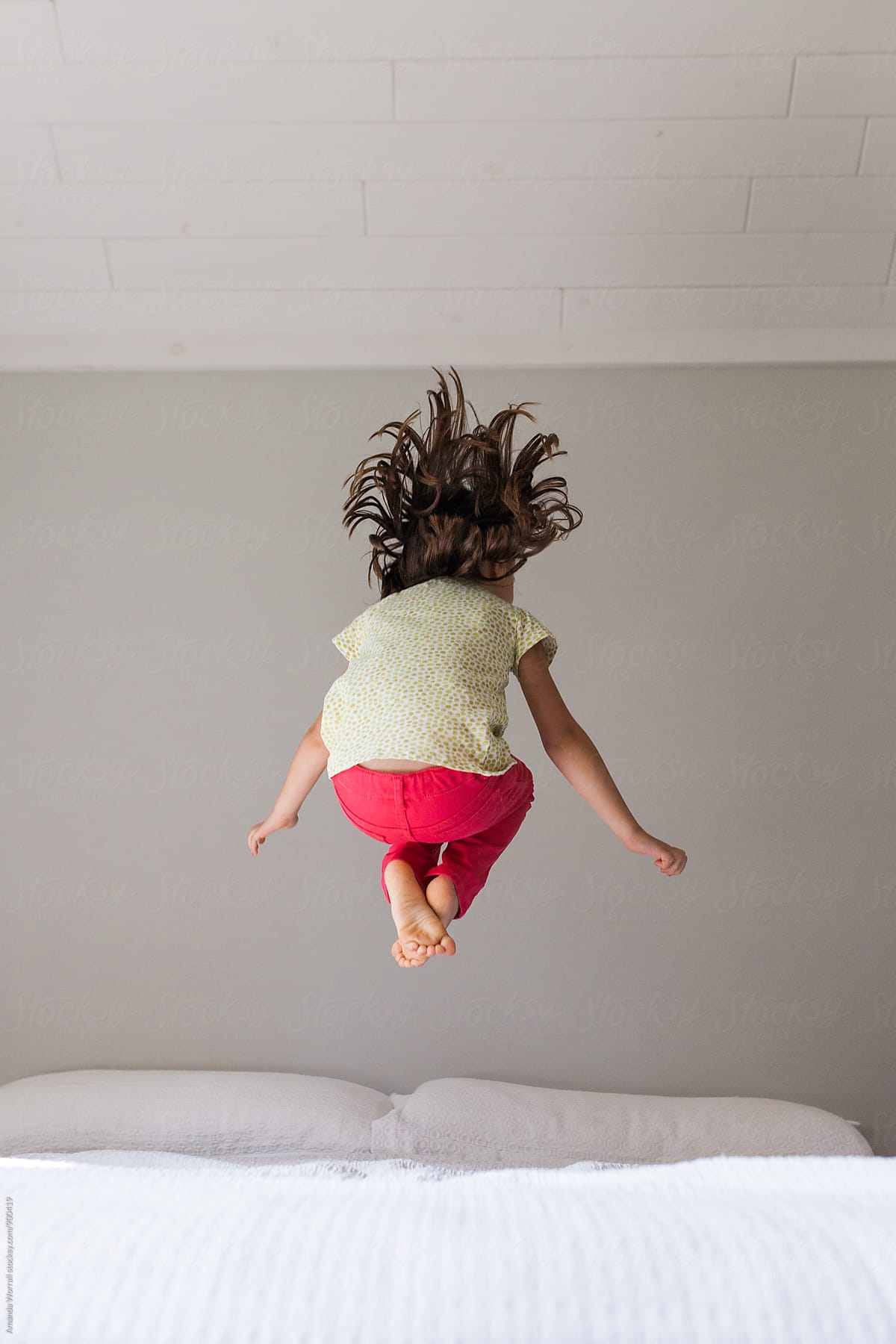 Unrecoginizable young girl wearing bright clothing jumping on a bed in a neutral bedroom