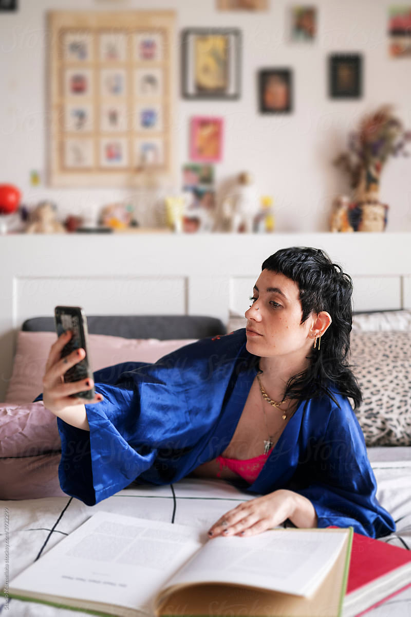 Young female in sleepwear taking selfie while reading book