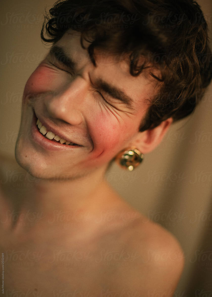 Portrait of a queer guy with curly hair and golden earrings who blushes and smiles broadly.