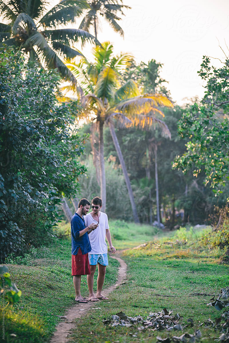 Two young men friends checking phone in a tropical land with palm trees and small dirt path. Kerala, India