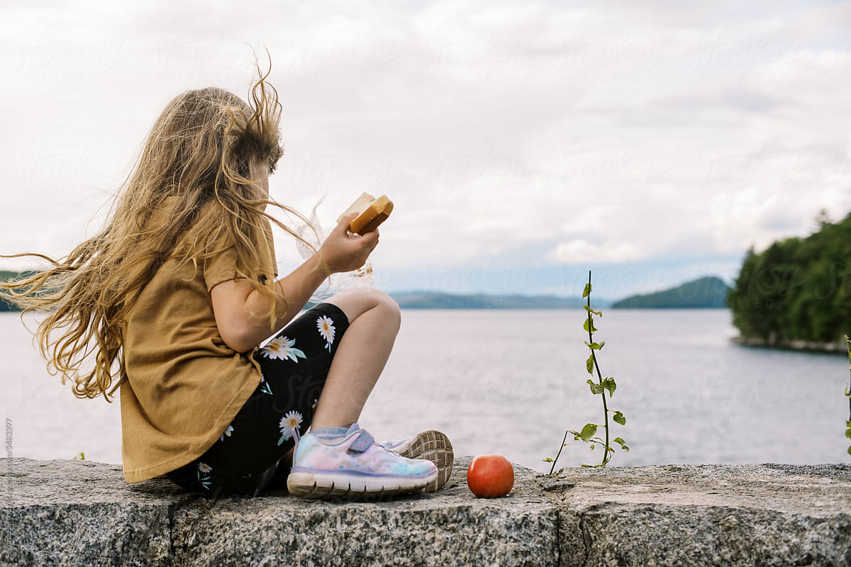 little girl sitting on rock wall overlooking a lake to eat lunch