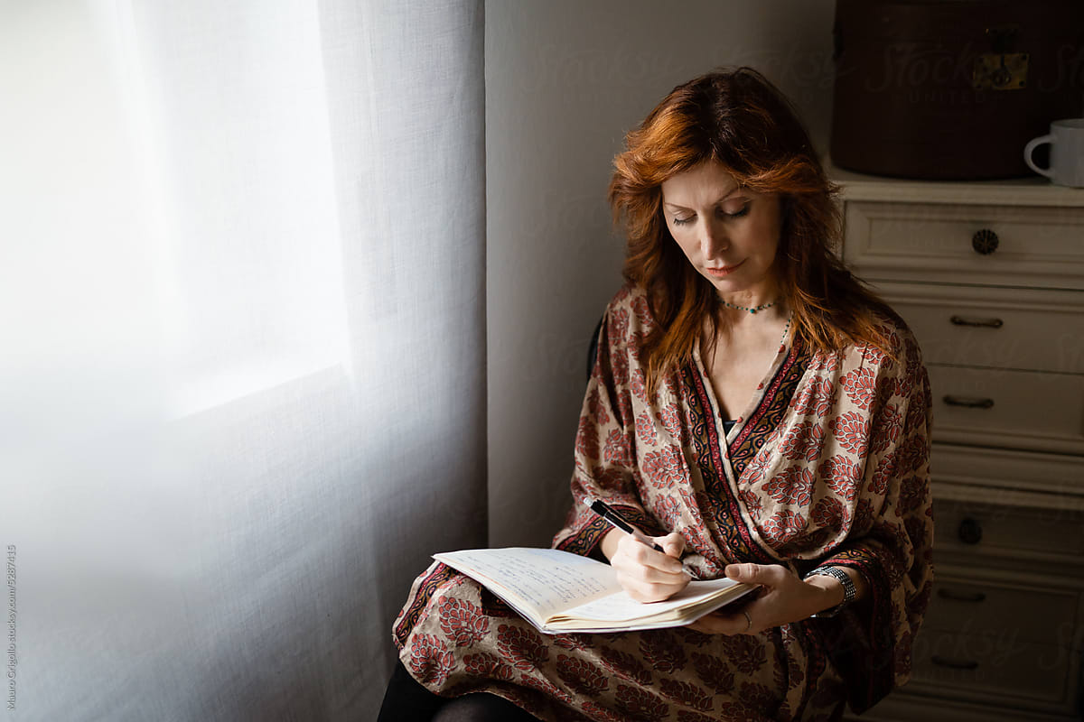 A woman writes in her notebook and sits near a window
