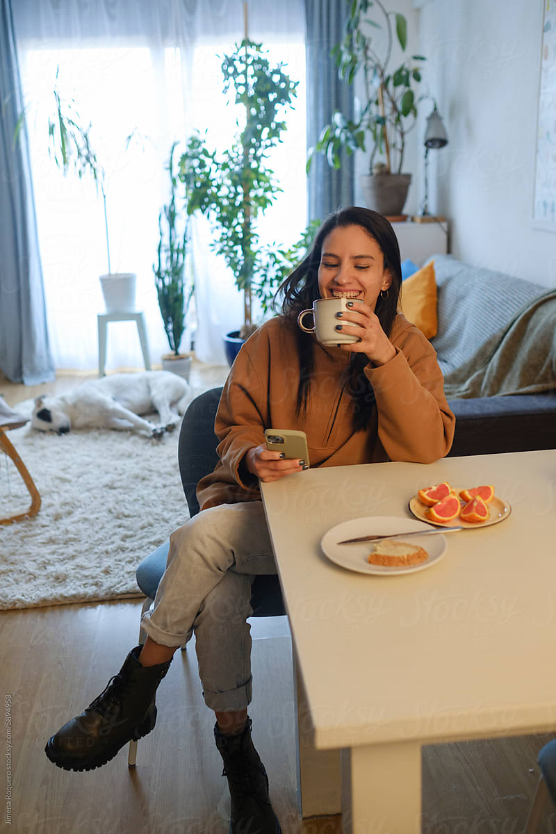 Woman at home having coffee while using smartphone smiling