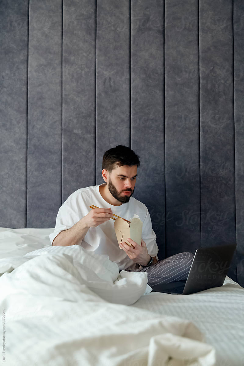 Young man eating noodles and using laptop on cozy bed